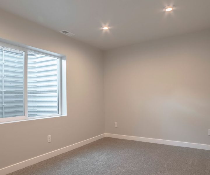 Clean, unfurnished basement room with beige carpet, light gray walls, white baseboards, recessed ceiling lights, and a single egress window with horizontal blinds—ready for customization or final staging.