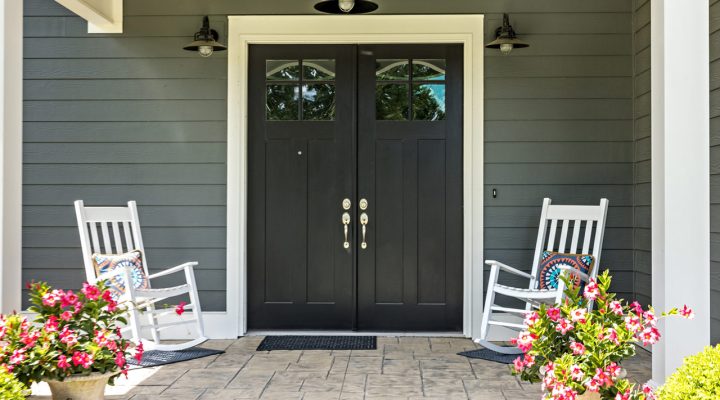Welcoming front entrance featuring charcoal-gray siding, black double doors with glass panels, white rocking chairs with colorful pillows, potted pink flowers, and a stamped concrete floor—showcasing curb appeal and exterior refinement.